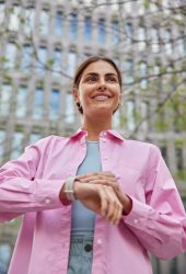 woman-smiles-gently-checks-time-wrist-watch-waits-someone-comes-meeting-wears-pink-shirt-poses-against-city-building_273609-55779